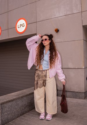 Joven con cabello largo y rizado, lleva chaqueta rosa, gafas de sol, pantalones anchos beige con detalles de encaje, sosteniendo una bolsa marrón cerca de señales de GLP.