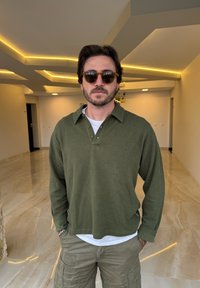 Polo a maniche lunghe ribbed verde oliva scuro con colletto bottonato, indossato sopra una t-shirt bianca, abbinato a pantaloni cargo chiari.