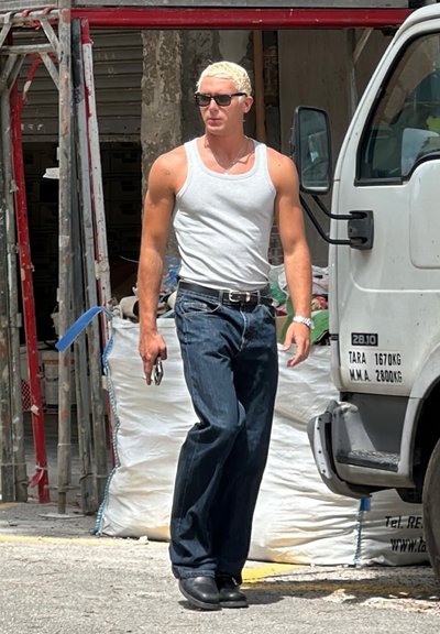 Camiseta sin mangas blanca, jeans azules y botas negras. Pelo rubio corto, gafas de sol y sosteniendo un smartphone. El fondo incluye materiales de construcción.