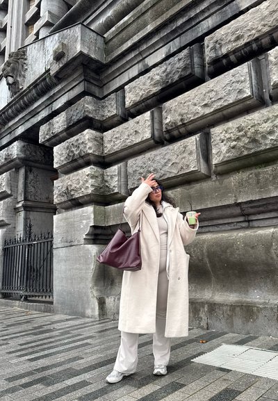 Largo abrigo beige sobre un conjunto beige, sosteniendo una bebida verde, de pie sobre un pavimento de piedra cerca de una pared gris texturizada. Un bolso burdeos cuelga de su hombro.