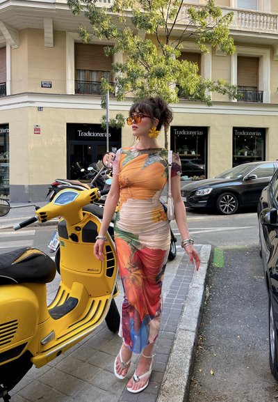 Vestido colorido con estampado floral, diseño ajustado, mangas cortas y una falda fluida. Los accesorios incluyen pendientes amarillos y gafas de sol.