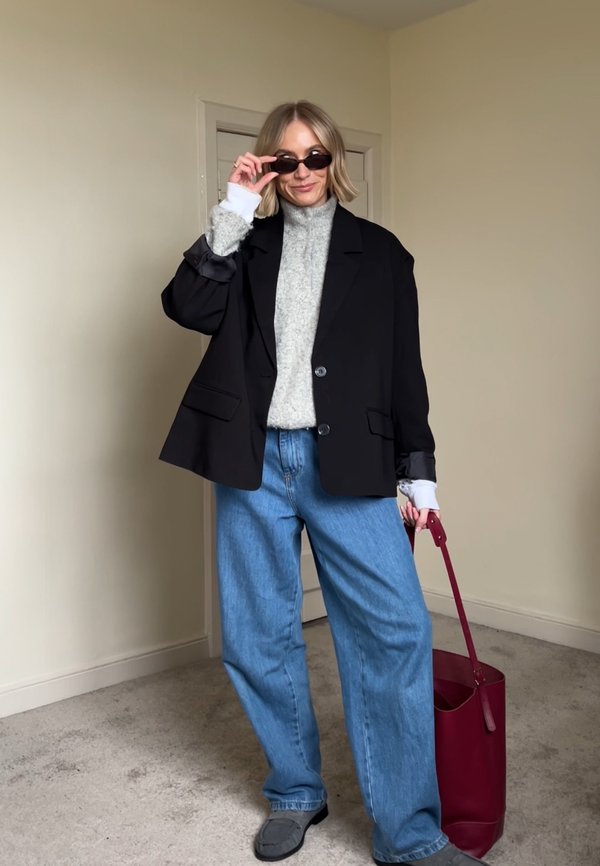 Femme ajustant des lunettes de soleil noires, portant un blazer noir sur un pull gris, un jean large bleu, des chaussures grises, et tenant un sac à main rouge dans une pièce neutre.