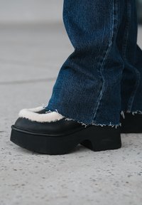 Chaussures compensées noires avec un bord en peluche crème, associées à un jean bleu effiloché. La chaussure présente un bout rond et une semelle épaisse.