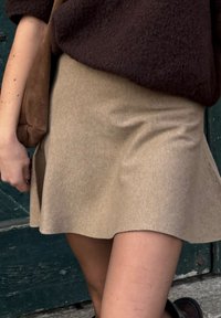 Jupe beige en velours côtelé coupe trapèze avec une texture subtile, tombant au-dessus du genou, associée à un pull marron foncé et un sac à bandoulière marron.