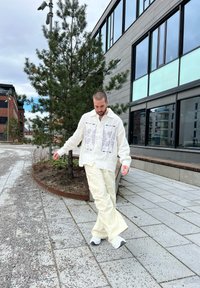 Vit broderad skjorta med blå mönster, krämfärgade cargo-byxor och vita sneakers. Utomhusmiljö med träd och modern byggnad.