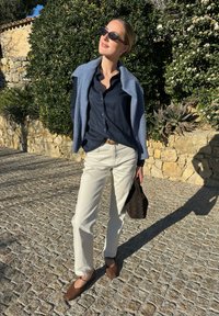 Το μοντέλο φορά μια ναυτική πουκάμισο με κουμπιά, μπεζ παντελόνια και καφέ επίπεδα παπούτσια. Μια ανοιχτού μπλε κάπα είναι ριγμένη στους ώμους της. Κρατά μια σκούρα πλεκτή τσάντα.