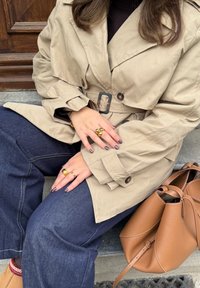 Béžový trench coat s opaskem v pase, tmavě modré široké džíny a hnědá kožená taška. Doplněno zlatými prsteny a vzorovanými nehty.