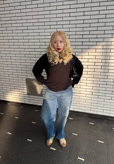 Top negro de manga larga combinado con unos vaqueros de pierna ancha de color azul claro. Los accesorios incluyen un pequeño bolso negro y zapatillas beige. Fondo de pared de ladrillo liso.