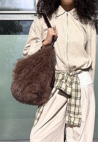 Individual in a beige zip-up jacket and pants, with a plaid shirt tied around their waist, carrying a large brown furry handbag over their shoulder.