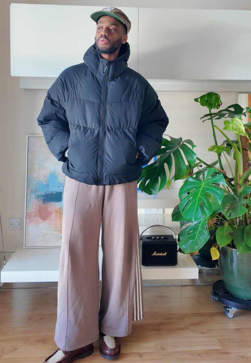 Mann in schwarzer Pufferjacke und beigen weit geschnittenen Hosen, der neben einer großen grünen Pflanze und einem kleinen schwarzen Marshall-Lautsprecher auf einem weißen Regal steht.