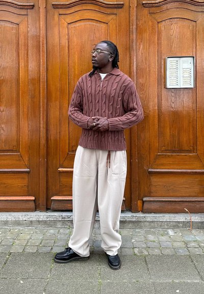 Hombre con gafas que lleva un suéter marrón de punto con trenzas y pantalones anchos beige claro se encuentra con las manos entrelazadas frente a grandes puertas de madera.
