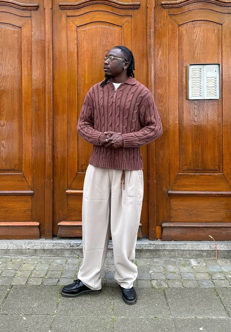 Uomo con occhiali che indossa un maglione marrone lavorato a trecce e pantaloni larghi beige chiaro, in piedi con le mani giunte davanti a grandi porte di legno.