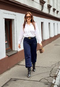 Femme portant une chemise blanche, un pantalon foncé, des bottes noires, des lunettes de soleil, tenant un petit sac à main marron en marchant sur le trottoir de la ville.