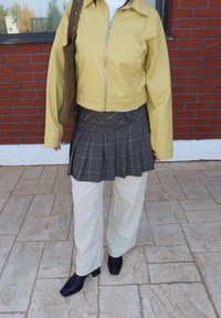 Persoon die een gele jas met ritssluiting draagt, een geplooide geruite rok over beige broeken, zwarte laarzen met hak, en een bruine schoudertas draagt.