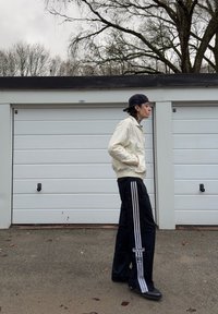 Light beige bomber jacket with a textured surface worn over black track pants featuring white stripes and the Adidas logo, paired with black shoes.