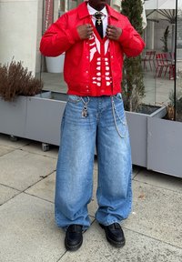 Casaco vermelho com fecho de correr sobre uma camisa branca, colete tricotado vermelho e branco, jeans oversized azuis claros e sapatos pretos com borlas. Acessórios metálicos incluídos.