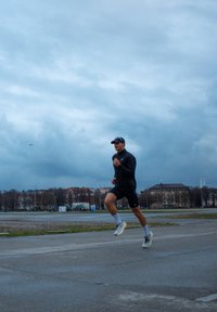 Vīriešu skrējējs melnā jakā un šortos, ar cepuri un baltām zeķēm, skrien pa pelēku virsmu zem mākoņainām debesīm.