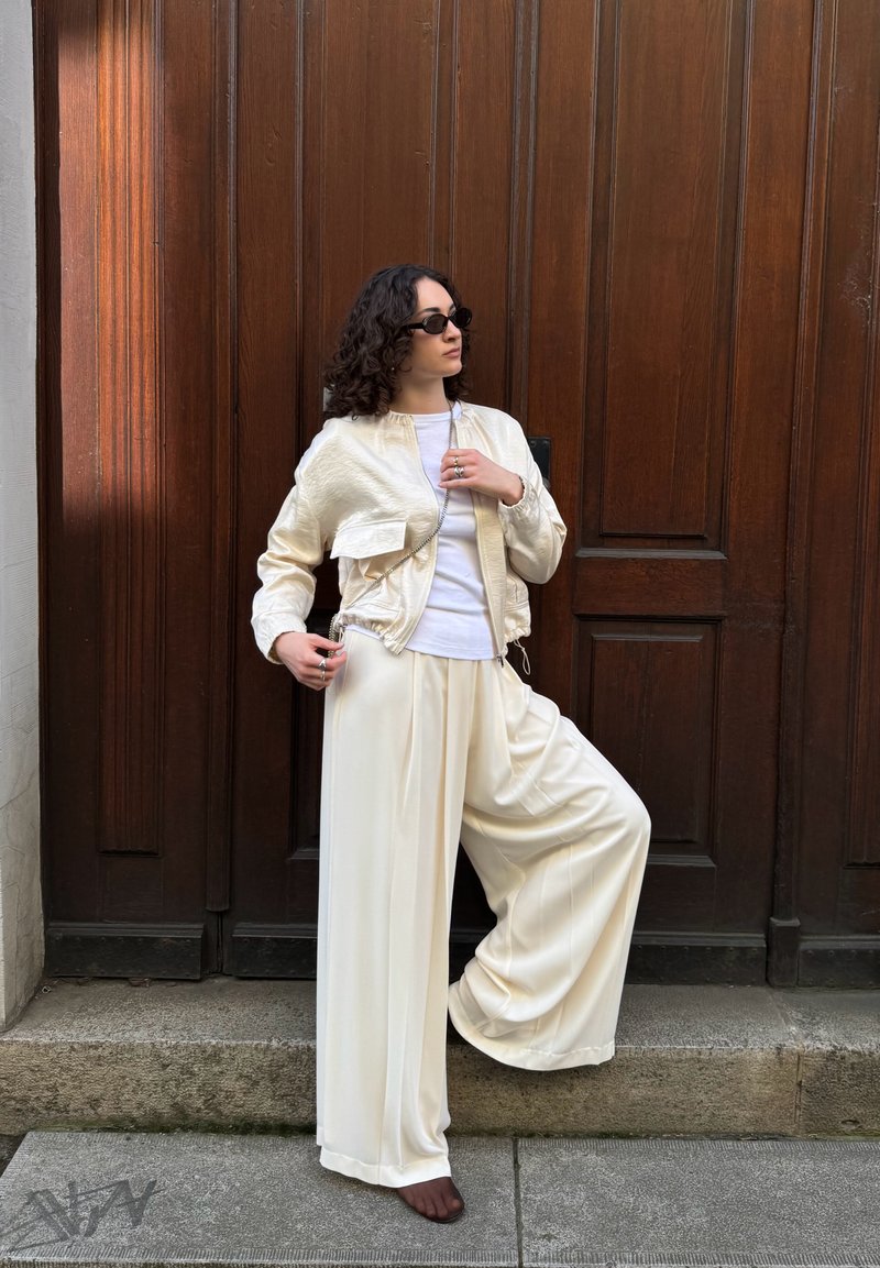 Personne aux cheveux bouclés portant des lunettes de soleil noires, une veste crème, une chemise blanche, un pantalon large crème, debout avec un pied sur une marche en pierre devant une porte en bois.