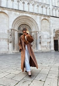 Brązowy płaszcz z sztucznego futra z szerokim kołnierzem, na podwójne guziki, z dużymi kieszeniami; w zestawieniu z jasnoniebieskimi dżinsami i czarnymi szpilkami.