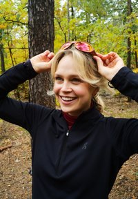 Juodas zip-up sportinis džemperis su tekstūru, tamsiai raudona polo megztinis po juo ir atspindinčios oranžinės saulės akiniai laikomi virš galvos.