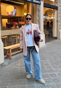 Sieviete, kas valkā saulesbrilles, beigu bleizeri, grafisku baltu T-kreklu, brīvus zilos džinsus un beigu kurpes, tur maroon krāsas somu, stāvējot pie kafijas veikala.