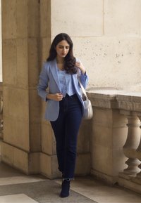 Lyseblå blazer over en matchende knapbluse, parret med mørkeblå jeans og ankelstøvler. En hvid skuldertaske bæres. Glatte teksturer.