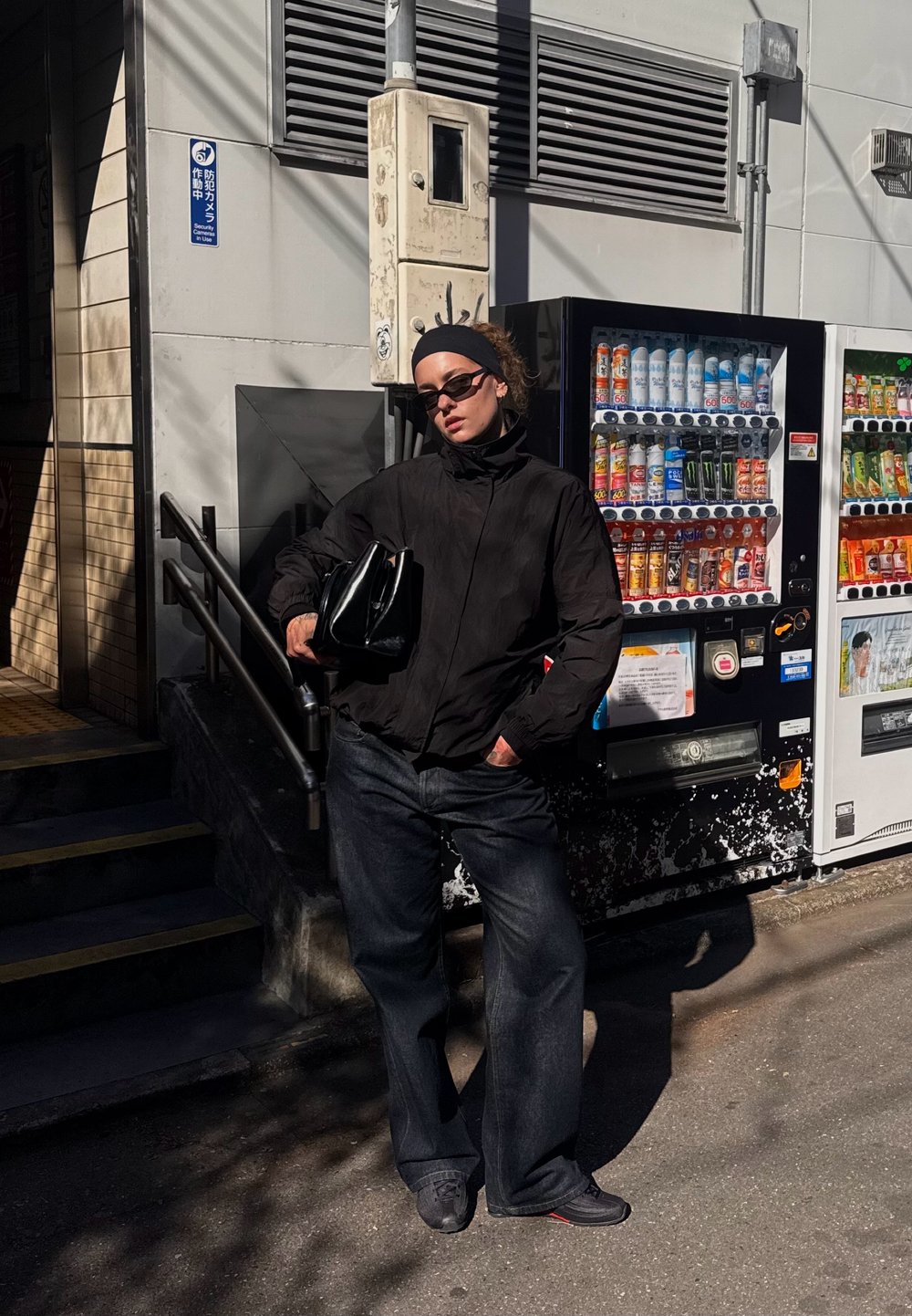 Jacket preto, jeans escuros e óculos de sol. A segurar uma mala preta. Em pé ao lado de máquinas de venda automática com várias bebidas. Cenário de rua iluminado pelo sol.