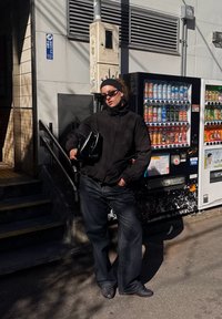 Jacket preto, jeans escuros e óculos de sol. A segurar uma mala preta. Em pé ao lado de máquinas de venda automática com várias bebidas. Cenário de rua iluminado pelo sol.