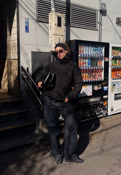 Chaqueta negra, jeans oscuros y gafas de sol. Sosteniendo un bolso negro. De pie junto a máquinas expendedoras con varias bebidas. Entorno de calle iluminada por el sol.