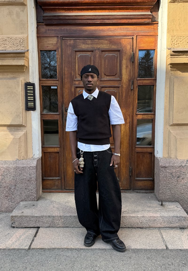 Mann iført svart baret, svart ermeløs genser over hvit skjorte, vide svarte bukser, står foran doble tredører.