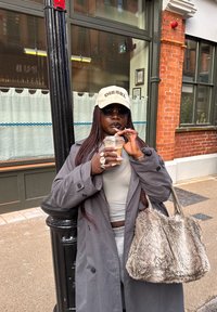 Mujer con el cabello trenzado, que lleva un abrigo gris y una gorra Diesel, bebe café con hielo mientras se apoya en un poste de luz negro en una calle de la ciudad.