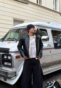 Fekete bőrdzseki krémszínű inggel, fekete, bő szárú nadrággal párosítva. A háttérben egy fehér, piros díszítésű kisbusz látható.