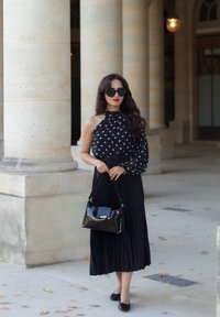 Top halter nero a pois bianchi con una manica trasparente, gonna nera plissettata, borsa nera lucida, occhiali da sole oversize neri, texture di tessuti lisci.