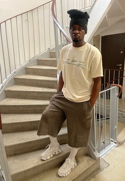 Camiseta beige claro con texto impreso, pantalones cortos marrones holgados, calcetines blancos y zapatillas deportivas grises. La persona está junto a una escalera de caracol.