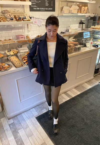 Abrigo navy de doble botonadura sobre una camiseta blanca de canalé, falda corta negra, medias negras transparentes, calcetines blancos y mocasines negros, en un ambiente de panadería.