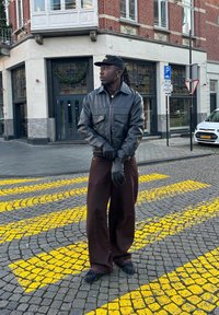 Homme en veste en cuir noire, gants et casquette, debout sur une rue pavée avec des marquages jaunes, devant un bâtiment en brique et une voiture garée.