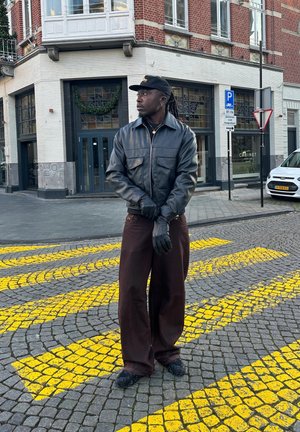 Hombre con chaqueta de cuero negra, guantes y gorra, de pie en una calle empedrada con marcaciones amarillas frente a un edificio de ladrillo y un coche estacionado.