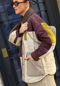 Femme portant une veste matelassée à blocs de couleurs marron, crème et jaune, avec un sac à chaîne, des lunettes de soleil écaille de tortue et des bouchons d'oreilles rayés.