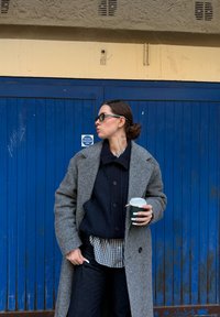 Femme portant un manteau gris, un pull noir et une chemise à carreaux tient une tasse de café, debout devant une porte métallique bleue avec un panneau d'avertissement.