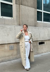 Beige trench coat nagy hajtókákkal, dupla gombolású, fehér ujjatlan felső és széles lábú fehér nadrág fölött viselve. Szürke sportcipők és napszemüveg a kézben.