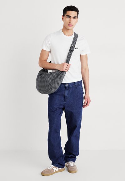 Bolso de hombro a cuadros grises, de tela suave, forma curvada, llevado por una persona con una camiseta blanca y jeans de mezclilla azul, zapatos de color claro.
