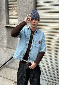 Chemise en denim surdimensionnée, pull marron en dessous, pantalon texturé foncé, et bonnet bleu à carreaux. Poche remarquablement visible sur la chemise.