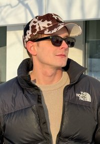 Homme portant une veste noire North Face, un pull beige, une casquette imprimée vache et des lunettes de soleil noires regarde sur le côté en extérieur sous le soleil.