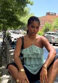 Jeune femme aux cheveux tressés portant un haut vert menthe à volants et un short noir, assise près d'un rack à vélos dans une rue ensoleillée de la ville.