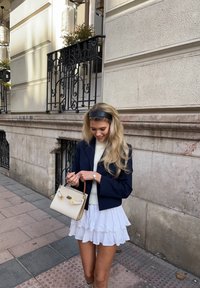 Ναυτικό μπλέιζερ, λευκή φούστα μίνι με ρίγες, μπεζ τσάντα χεριού με χρυσές λεπτομέρειες, μαύρη κορδέλα για το κεφάλι και ρολόϊ. Αστική τοποθεσία με πέτρινους τοίχους.