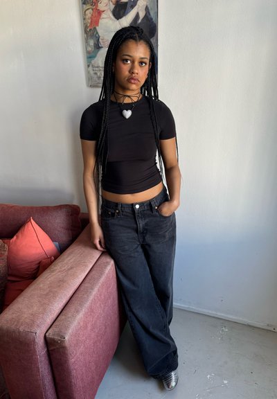 Top corto negro de manga corta, jeans negros de pierna ancha de tiro alto y zapatos de tacón metálicos; la modelo está de pie al lado de un sofá rosa con cojines.