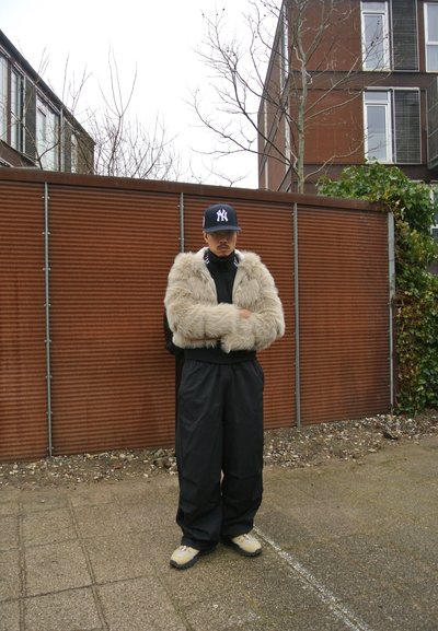 Chaqueta corta de piel sintética en crema, suéter de cuello alto negro, pantalones anchos negros, zapatillas oscuras y una gorra de béisbol azul marino con logotipo blanco.