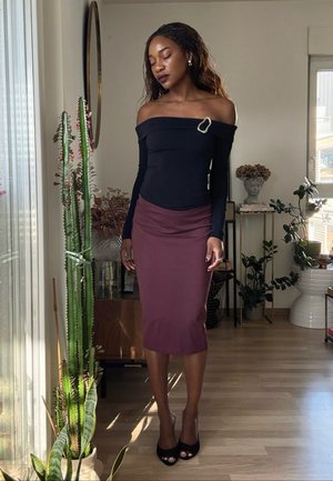 Mujer con el cabello largo trenzado, vestida con una blusa negra de hombros descubiertos y una falda marrón, de pie descalza en una habitación iluminada por el sol con plantas altas.