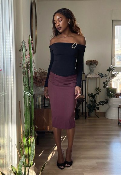 Mujer con el cabello largo trenzado, vestida con una blusa negra de hombros descubiertos y una falda marrón, de pie descalza en una habitación iluminada por el sol con plantas altas.