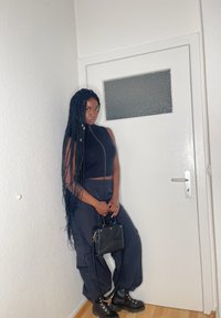Schwarzes gestricktes Cropped-Top mit Reißverschluss, hochtaillierte marineblaue Cargo-Hosen, schwarze Stiefeletten und eine kleine Handtasche, posierend vor einer weißen Tür.